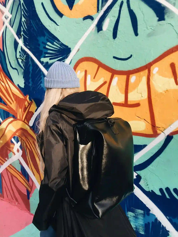 Person wearing a black leather backpack with a vintage design, styled for everyday use, against a colorful graffiti wall.